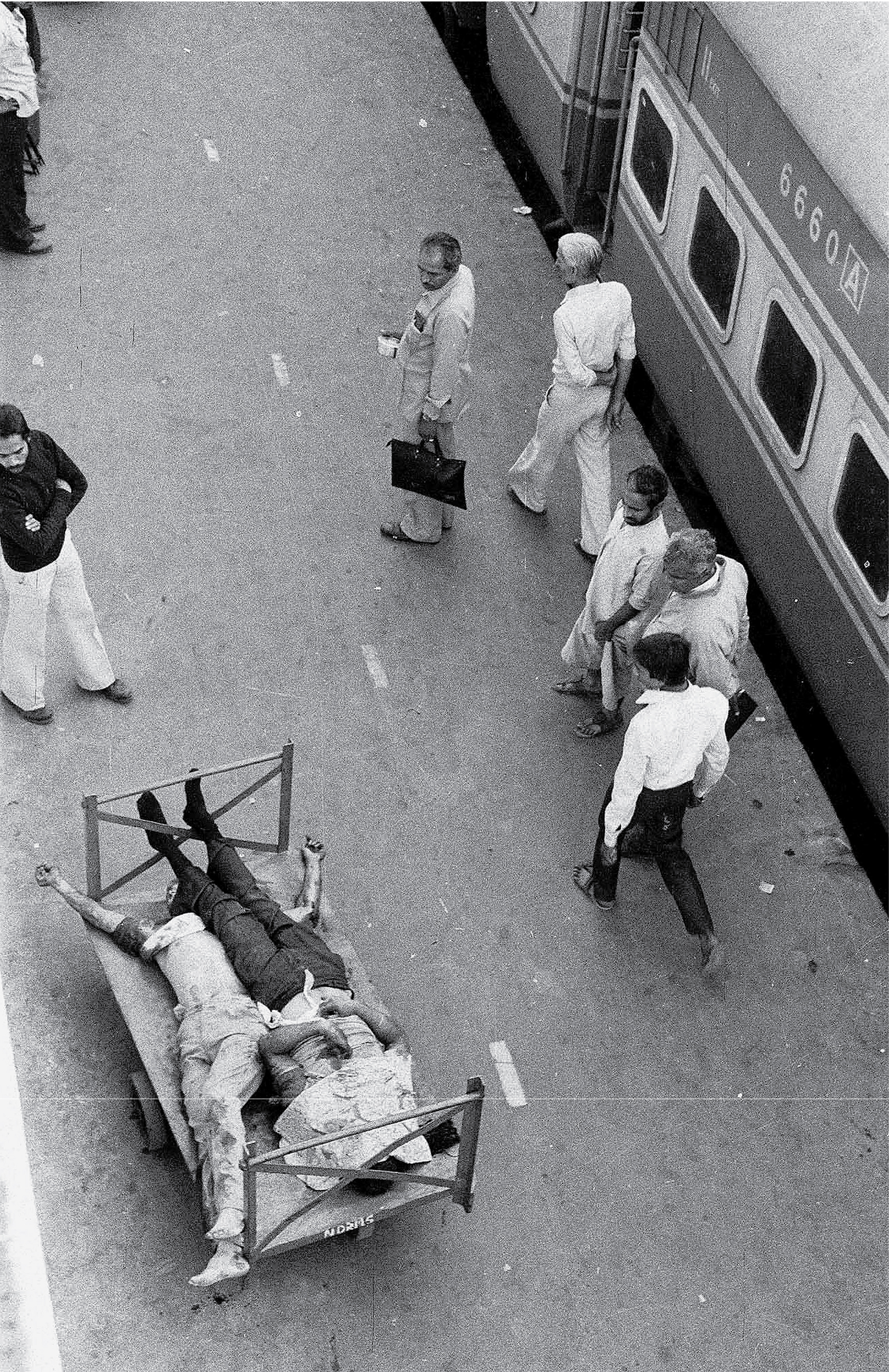 Commuters walk past two bodies on a parked luggage trolley on the platform at New Delhi Railway Station, November 1984 | Image credit: Ashok Vahie Commuters walk past two bodies on a parked luggage trolley on the platform at New Delhi Railway Station, November 1984 | Image credit: Ashok Vahie